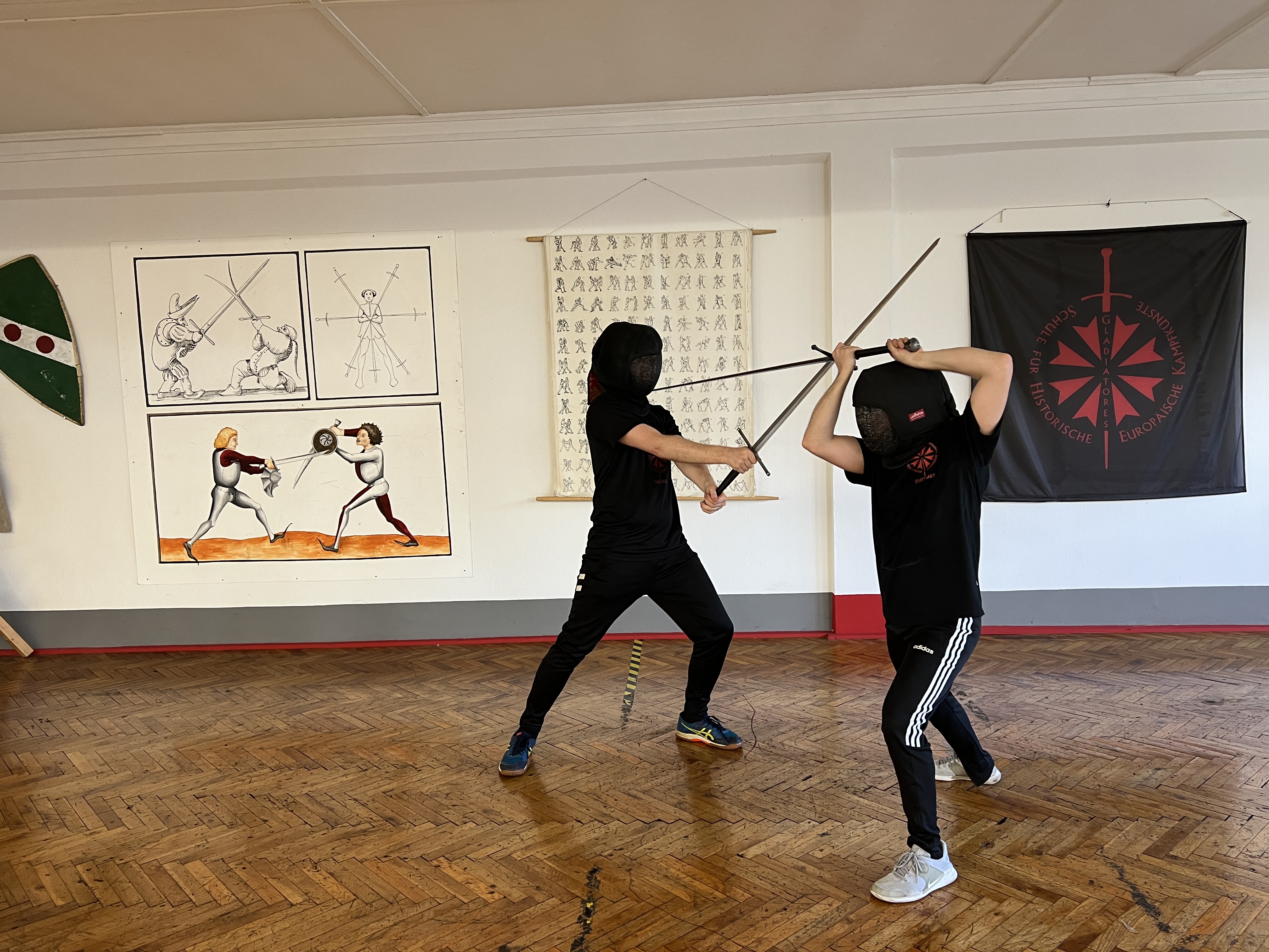 Zwei Fechtende im Training mit dem Langen Schwert vor Fechtbuch-Tafeln und dem Gladiatores-Banner
